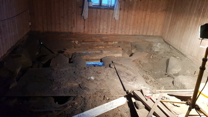 Rivet golv i gammal drängstuga där bara jorden syns, med hål ner till en underliggande jordkällare och verktyg synliga.