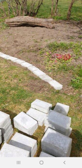 Stenplattor staplade inför anläggning av trädgårdsgång med gräs, jord och blommor i bakgrunden.