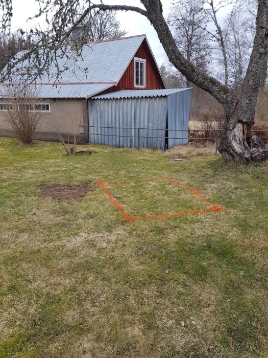 Planerad yta för sandlåda markerad med orange spray på gräs framför ett röd-vitt lantligt uthus, med en stor trädgren överst.