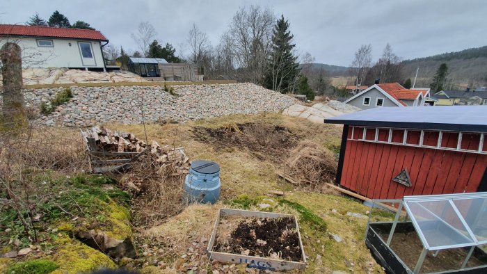 Trädgård som sluttar med övervuxna nässlor, stenmur, röd stuga och växthus.