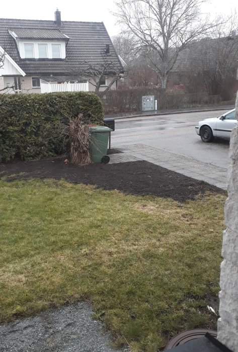 Område med nyligen jämnad jord bredvid gräsmatta och stenplattor under gröna soptunnor, tidigare plats för häck.