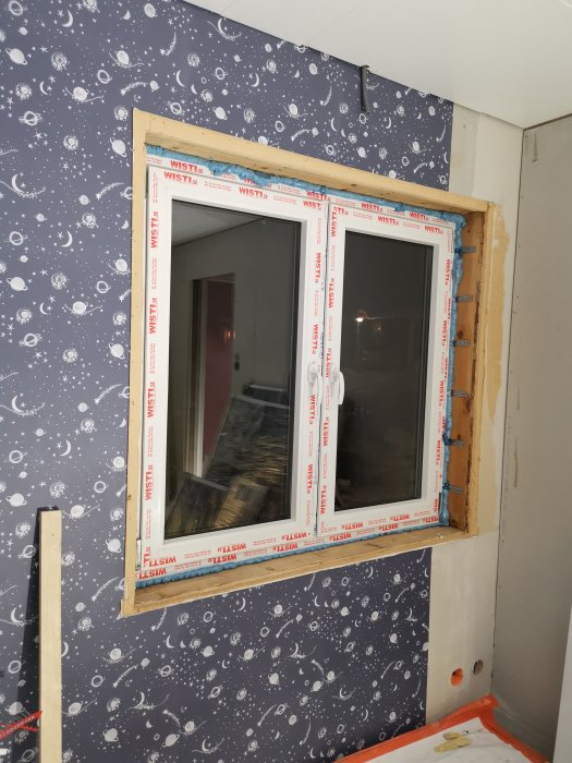 Nyligen installerat fönster i vägg med rymdmönstrad tapet under renovering.