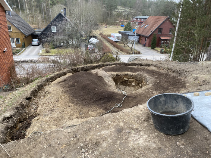 Jordarbete i trädgård med upphögd kant och en hög mörk jord, hink i förgrunden, utsikt över bostadshus.