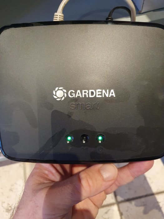 Hand håller en Gardena Smart Gateway med två gröna lysdioder tända, vilket indikerar ström och wifi-anslutning.