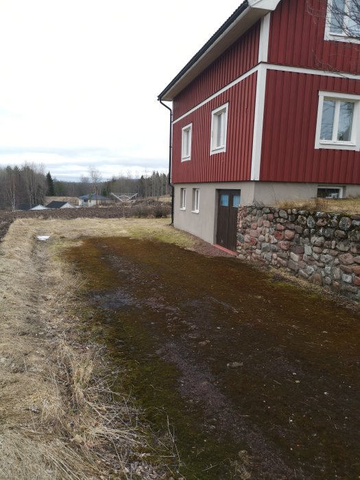 Mossbelagd yta framför rött hus med vit kant och stenfot, grå himmel och bar mark.