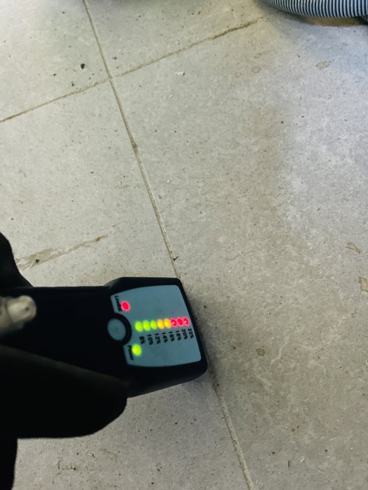 Fuktindikator i en hand visar höga fuktighetsnivåer på ett stengolv med mörkare fogar.