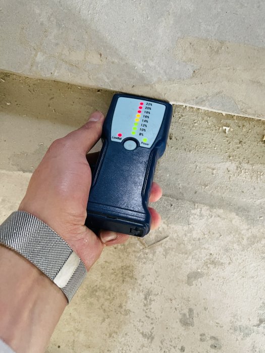 Hand håller en fuktmätare mot ett betonggolv med höga fuktindikeringar efter en vattenläcka i huset.