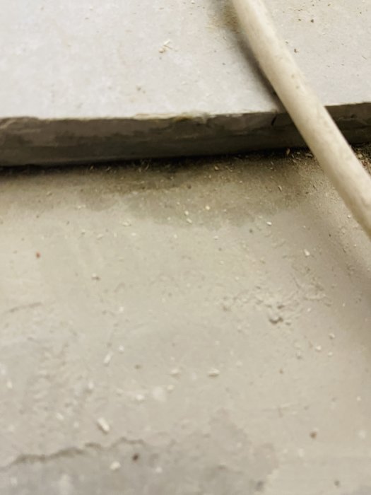 Närbild på en betonggolv med mörkare fogar och en sladd, indikativt för en tidigare vattenläcka och ihållande fuktproblem.