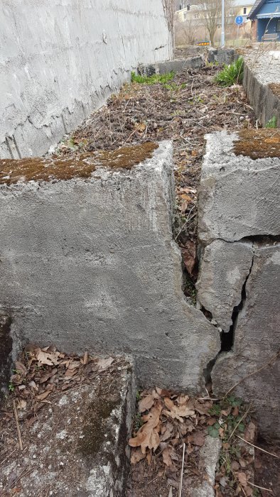 Äldre stödmur med synliga sprickor och lutning, omgiven av torra löv och vegetation, vilket indikerar brist på dränering.