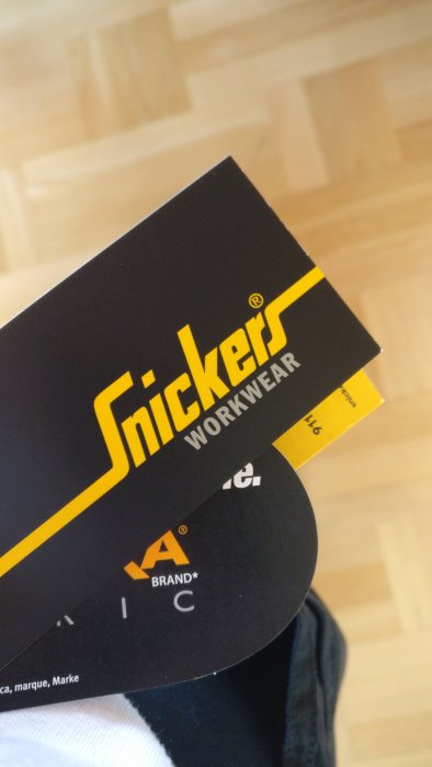 Etikett från Snickers Workwear arbetskläder som hålls av någon i förgrunden, med suddig bakgrund av ett trägolv.