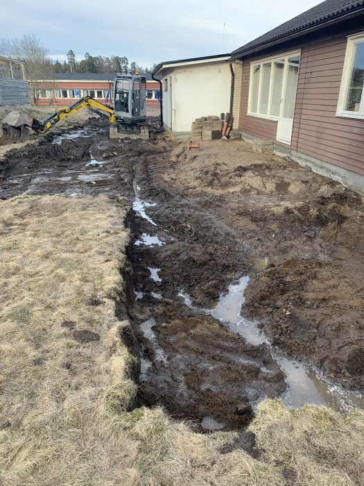 Grävd plats för altanbygge med blöt jord och en grävskopa intill ett hus.