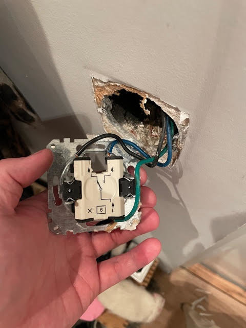 Öppen eldosa med synliga ledningar och en strömbrytare som hålls i en hand framför en trasig vägg.