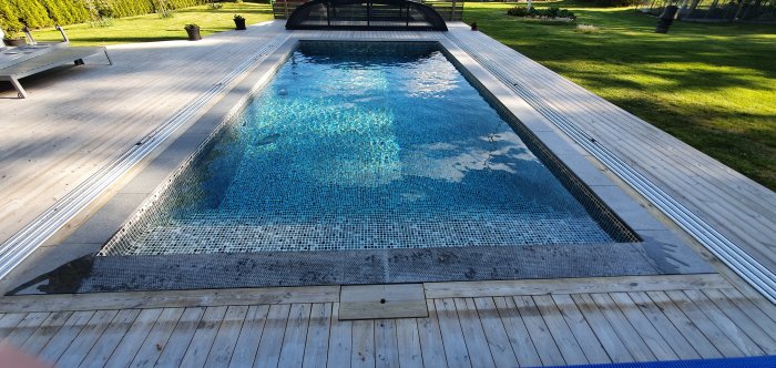 Pool med svart aluminiumprofil och öppet tak två meter bort, omgiven av trätrall och grön gräsmatta.