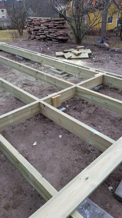 Pågående konstruktion av trädäck med träreglar placerade på jordad mark, med högar av virke i bakgrunden och gulaktigt hus.