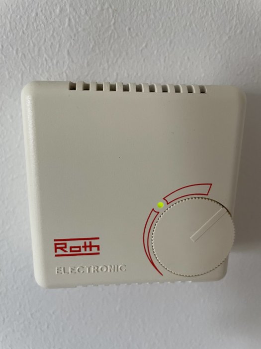 En Roth Electronic rumstermostat utan display monterad på en vit vägg, med en grön lysdiod tänd.