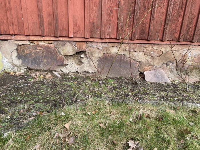 Äldre stengrund med synliga sprickor och skador under en rödmålad trävägg, gräs framför.