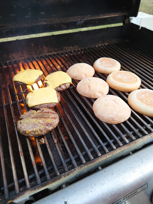 Hamburgare och bröd grillas på en gasolgrill med synliga lågor.