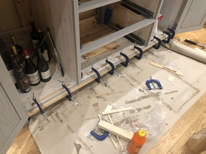 Köksskåp under konstruktion med trevägstvingar, träbitar, lim och vinflaskor.