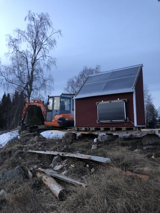 Torkhus med solpaneler står bredvid en grävmaskin och virke på en byggarbetsplats under tidig kväll.