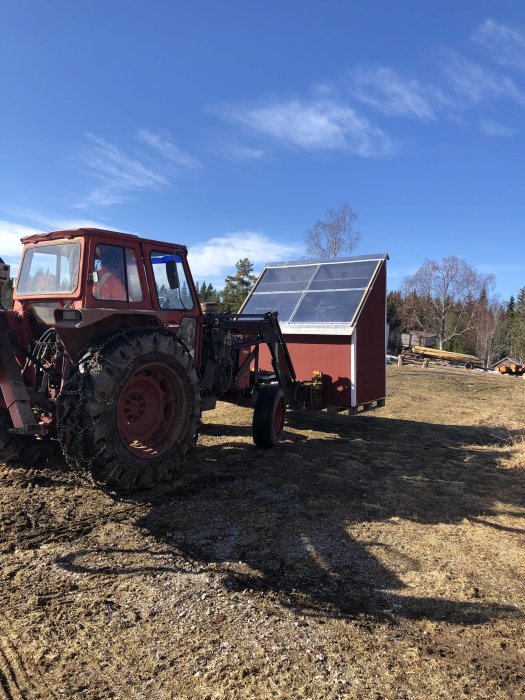 Traktor utomhus bredvid en liten röd bod med solpaneler under en klarblå himmel.