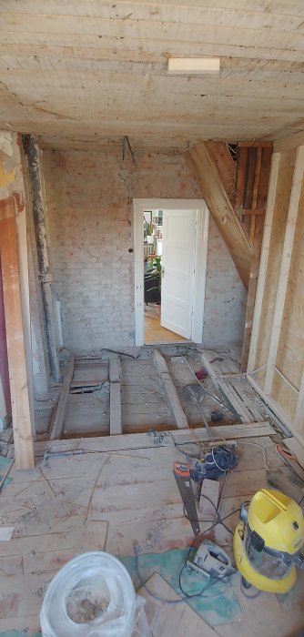 Rivningsfas av ett badrum med synligt bjälklag, verktyg på golvet och en öppen dörr mot ett annat rum.