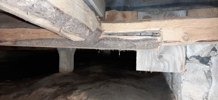 En skadad träbalk med en glipa där några spikar är synliga i en krypgrund under ett hus.