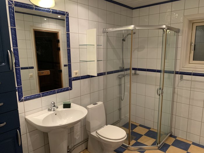 Renoverat badrum med vita kakelväggar, blå dekorremsor, duschhörna, handfat och toalett.