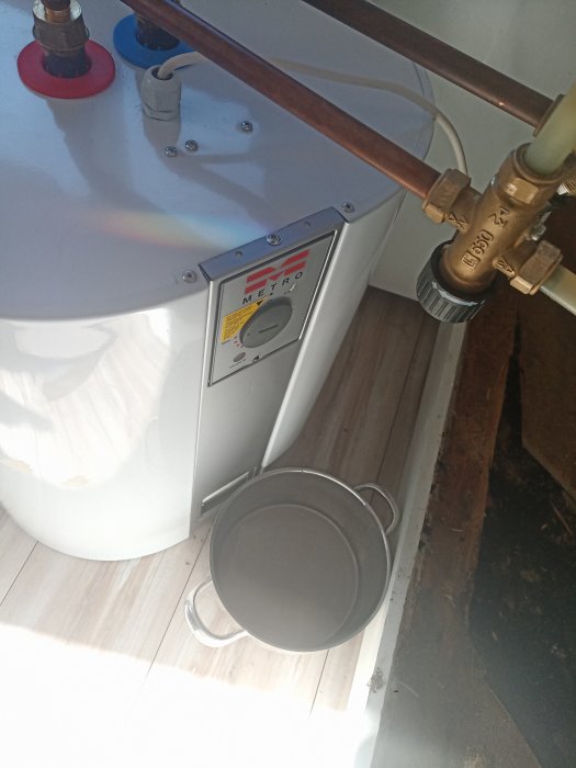 Varmvattenberedare i ett hem med justeringsknapp, kopparledningar och säkerhetsventil, bredvid en grå hink.