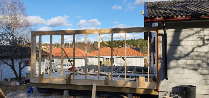 Nybyggd trävägg på stolpar med grannhus och träd i bakgrunden.