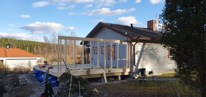 En nybyggd väggstomme av trä intill ett hus med verktyg och material synliga i förgrunden, i ett soligt bostadsområde.