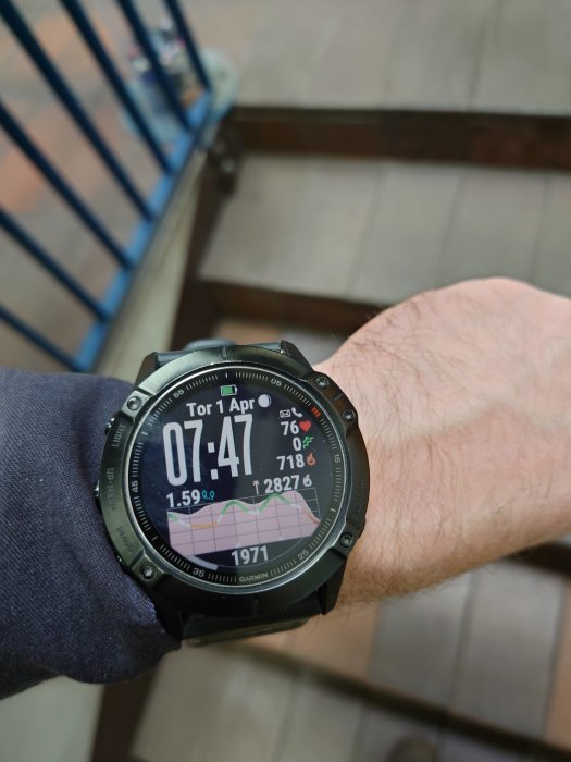 Smartklocka på en persons handled visar tid, datum, och aktivitetsdata mot en trappbakgrund.