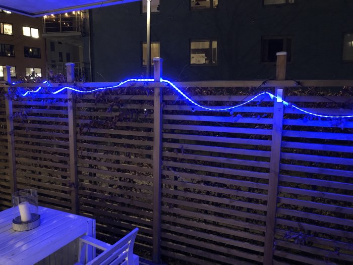 Blå LED-ljusslinga upphängd på ett trästaket med byggnader i bakgrunden på kvällen.