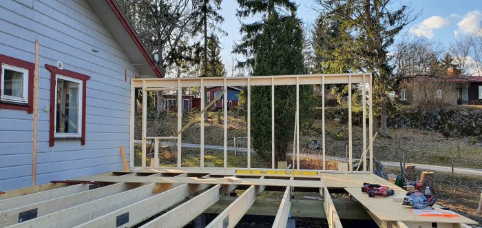 Nybyggd trävägg med reglar och stolpar på en husgrund, i ett bostadsområde under solig himmel.