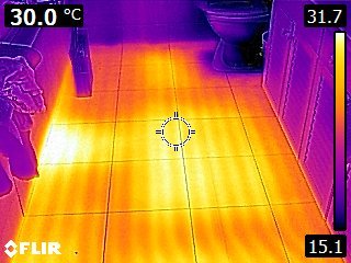 Termisk bild av golvvärme i badrum, kablar synliga vid toalett, spegelbild av person, FLIR-kamera.