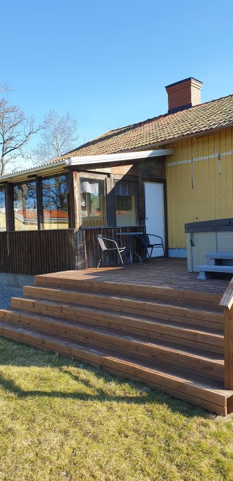 Trädäck med trätrappa framför ett gult hus med inglasad veranda och utemöbler under en klarblå himmel.