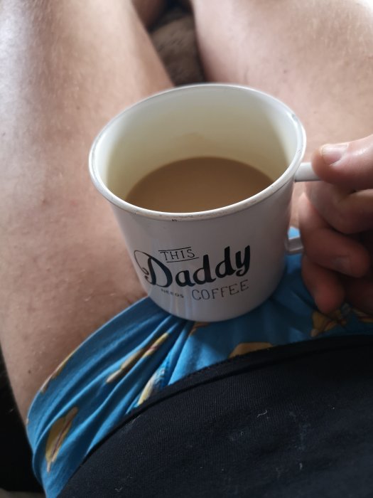 Person håller en kaffemugg där det står "THIS DADDY NEEDS COFFEE" med korsade ben i bakgrunden.