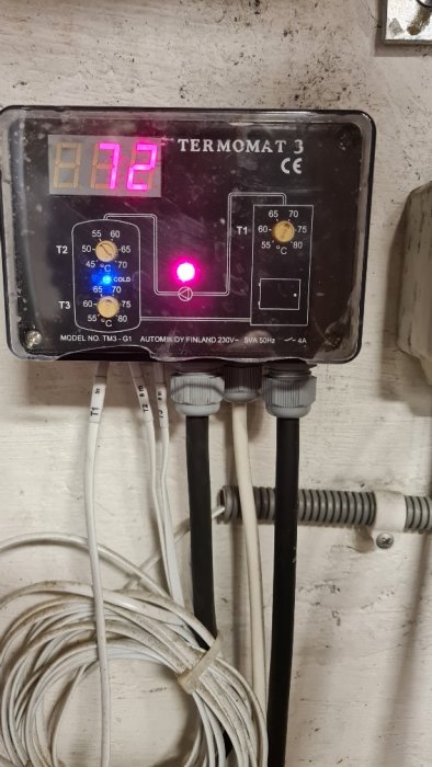 Digital termostatpanel med blå och röd lampa tända, omgiven av kablar och röranslutningar.