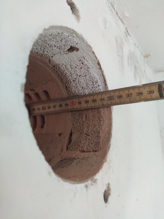 Mätning av ett ventilationshål i en vägg med ett måttband som visar diameter, med lager av tegel och puts.