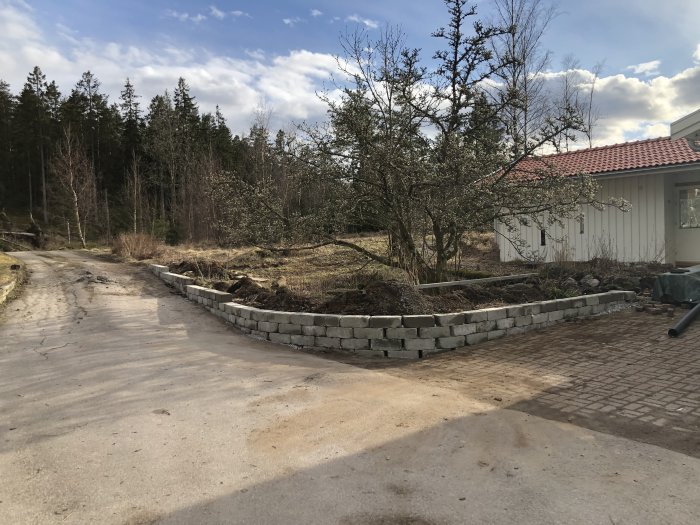 Nybyggd stödmur av gråa betongblock i en trädgård med grusväg och träd i bakgrunden.