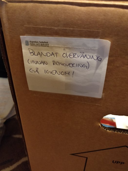 Etikett med texten "BLANDAT ÖVERVÅNING (inuti Renovering) GÅ IGENOM!" på en brun pappkartong.