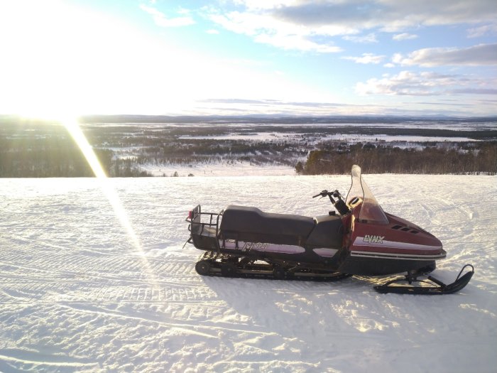 Röd snöskoter parkerad på snötäckt backe med solnedgång och vidsträckt landskap i bakgrunden.