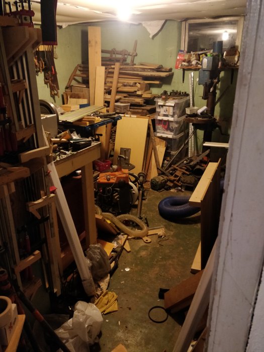 Hög med virke och verktyg i ett rörigt garage, spån och redskap på golvet.
