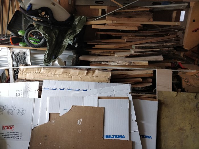 Hög med diverse trämaterial och kartonger i en rörig garageverkstad.