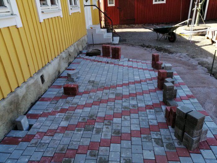 Nybyggd uteplats med röda och grå markstenar vid gult hus, ofärdig med staplar av stenar.