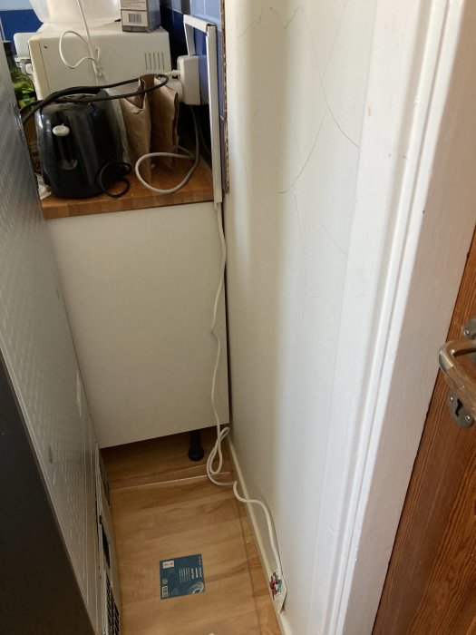 Elektrisk kabel löper slarvigt längs golvet och väggen bakom ett skåp med hushållsartiklar.