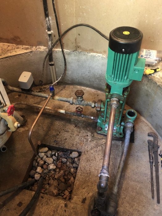 En grön industrivattenpump placerad nära en vägg med synliga rör och stenar på golvet.