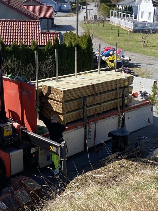 Leverans av trallvirke på lastbilsflak i en uppfart med arbetare och villaområde i bakgrunden.