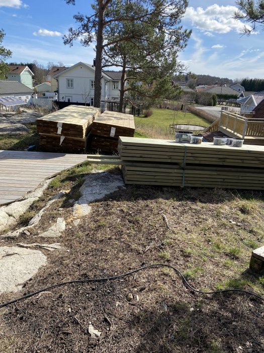 Trallvirke och byggmaterial staplade utomhus bredvid en träaltan, med en villa i bakgrunden.