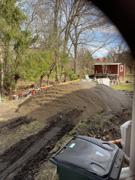 En hög med grus för motfyllnad nära en poolplats med byggmaterial spridda, tagen genom ett fönster.