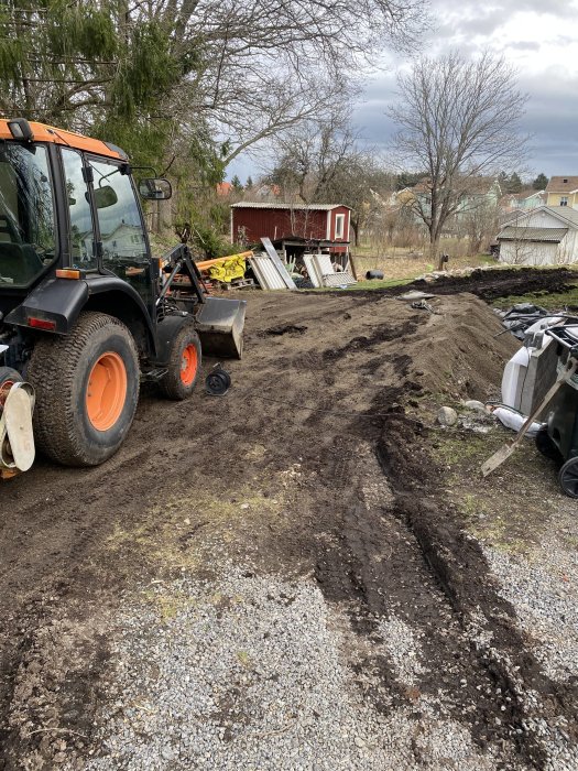 Traktor på en blivande byggarbetsplats med jordhögar, grus och byggmaterial i bakgrunden.